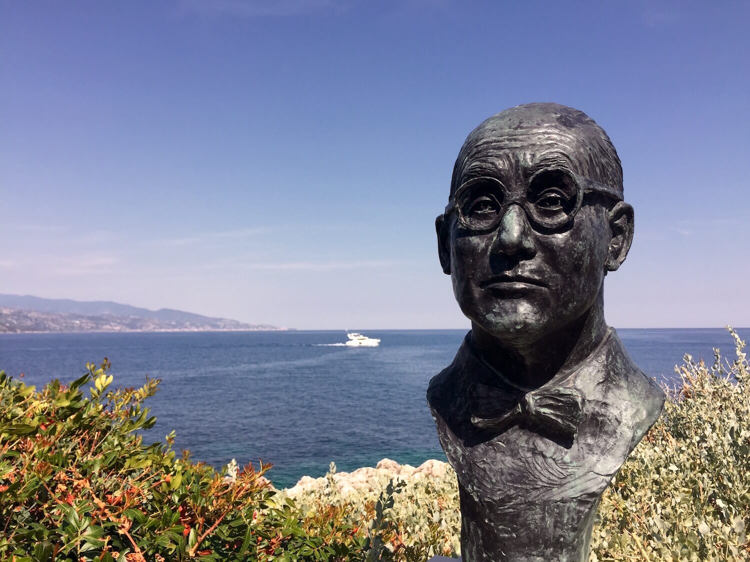 Corbusier Statue overlooking blue sea with yacht cruising along