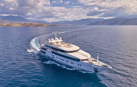 Aerial view of the charter yacht Marquise cruising through deep blue sea, leaving a curved white wake behind, with rugged mountains and a partly cloudy sky stretching across the horizon.